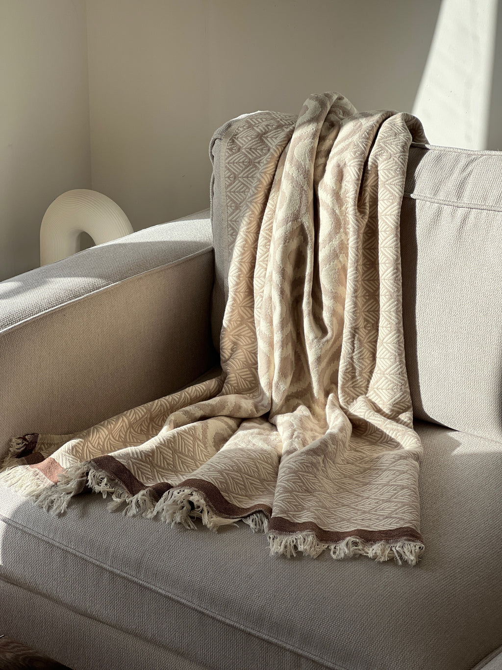 Cozy beige patterned throw blanket with tassels on a light sofa, perfect for cozy living spaces.